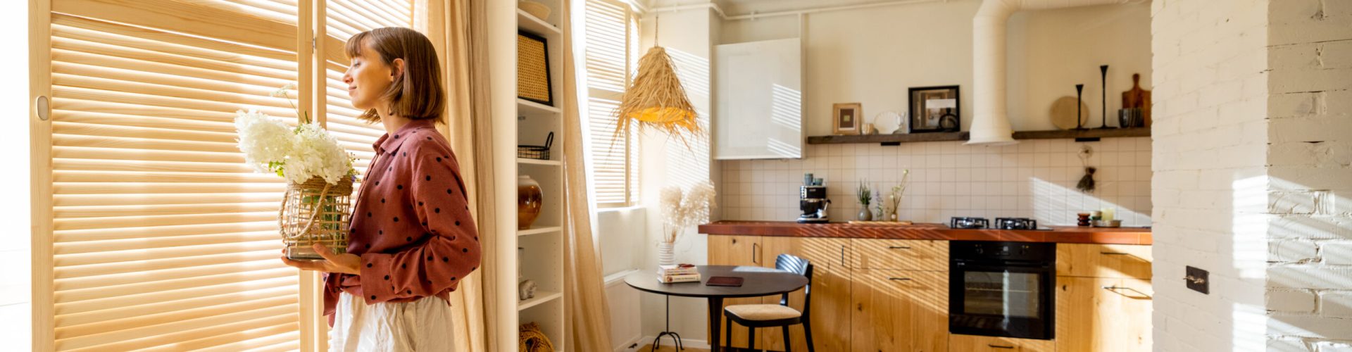 Woman walks with a vase of flowers, decoration and having some house chores at home. Wide interior view on sunny studio apartment in beige tones